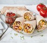 Stack of veggie burritos on a place next to a glass pot of chopped tomatoes